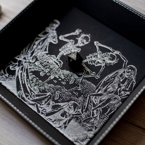 K&ouml;nnte beinhalten: Schwarzes Leder-W&uuml;rfeltablett mit einem wei&szlig;en Design von tanzenden Skeletten. Ein schwarzer 20-seitiger W&uuml;rfel befindet sich im Tablett.