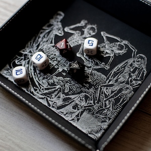 K&ouml;nnte beinhalten: Ein schwarzes Leder-W&uuml;rfeltablett mit einem wei&szlig;en Skelett-Design und sechs W&uuml;rfeln. Die W&uuml;rfel sind wei&szlig; mit blauen Zahlen und einem schwarzen W20.