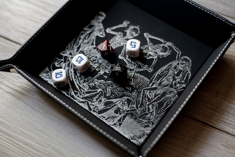 K&ouml;nnte beinhalten: Ein schwarzes Leder-W&uuml;rfeltablett mit einem wei&szlig;en Skelett-Design und sechs W&uuml;rfeln. Die W&uuml;rfel sind wei&szlig; mit blauen Zahlen und einem schwarzen W20.