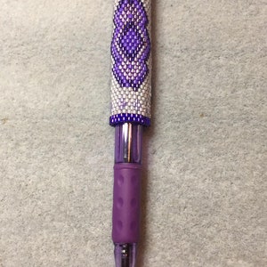 May include: A purple pen with a white and purple beaded design. The beads are arranged in a geometric pattern.