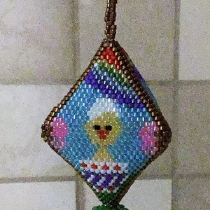 Peut inclure: Une décoration en perles en forme de diamant avec un motif arc-en-ciel coloré et un poussin jaune avec un chapeau blanc au centre. L'ornement est suspendu à un crochet blanc.