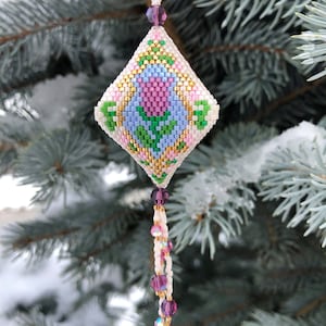 Könnte beinhalten: Ein weißer, rosa, blauer, grüner und goldener Perlen-Anhänger in Diamantform mit einem floralen Design. Der Anhänger hängt an einem Zweig einer Blaufichte.