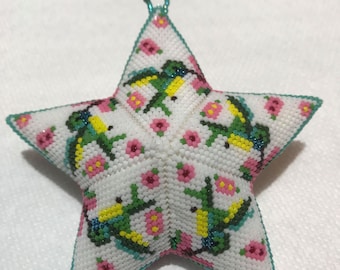 May include: A white star-shaped ornament with a beaded design featuring pink flowers, green leaves, and blue and yellow birds. The ornament has a loop at the top for hanging.