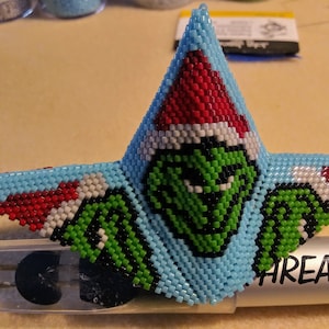 May include: A blue, green, red, and white beaded Christmas ornament shaped like a star with a Grinch face in the center. The ornament is sitting on a white surface.