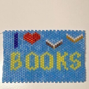 Puede incluir: Un llavero de cuentas azules con las palabras "Me encantan los libros" en cuentas amarillas. El "amor" está representado por una cuenta de corazón roja.