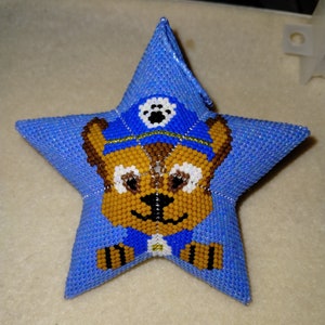 Op de afbeelding: Een blauwe stervormige ornament met een kralenbeeld van een cartoonhond die een blauwe en gele politiehoed draagt. De hond heeft bruine vacht, witte ogen en een zwarte neus.