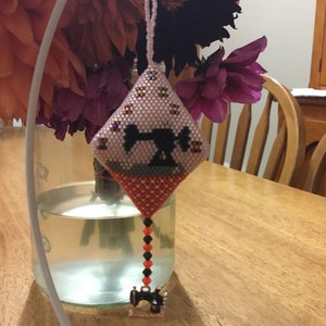 May include: A pink and orange beaded ornament shaped like a diamond with a black sewing machine design in the centre. The ornament is hanging from a white stand.