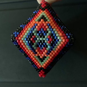 May include: A diamond-shaped beaded ornament with a geometric pattern in shades of red, orange, blue, black, and green.