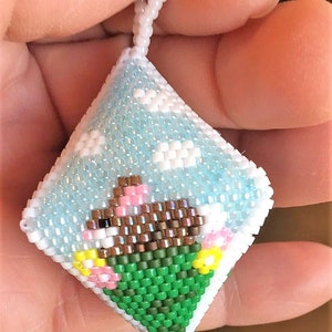 Könnte beinhalten: Ein kleiner, quadratischer Kissenanhänger aus Perlen mit einem braunen Bären, der eine rosa Blume auf grünem Hintergrund trägt. Der Hintergrund ist hellblau mit weißen Wolken.