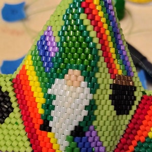 May include: A green, rainbow, and white beaded gnome ornament. The gnome is wearing a green hat and a white beard. The ornament is made of small, colorful beads.
