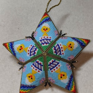 May include: A star-shaped ornament made of colorful beads, featuring a repeating pattern of yellow chicks in blue and white egg shells. The ornament has a gold loop for hanging.