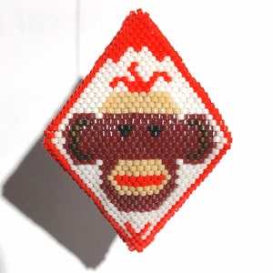 Puede incluir: Adorno con forma de diamante con un diseño de cara de mono con cuentas. El mono tiene una cara roja, gafas de sol y una boca beige. El borde es rojo y blanco, con fondo blanco.