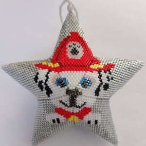 May include: A gray star-shaped ornament with a beaded design of a dog wearing a red firefighter hat. The dog has white fur, black spots, blue eyes, and a red bow tie.