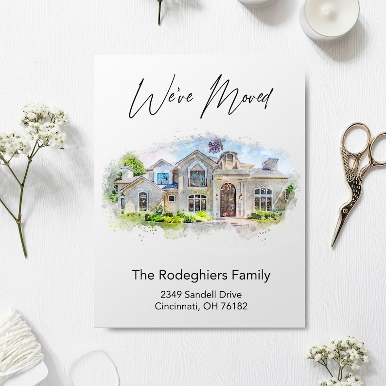 A personalized "We’ve Moved" announcement card featuring a custom watercolor illustration of a large, two-story stone house. The card is displayed on a white wood surface surrounded by aesthetic props including white baby's breath flowers, a spool of twine, a tealight candle, and ornate gold scissors. The text on the card reads "The Rodeghiers Family" with a Cincinnati, Ohio address.