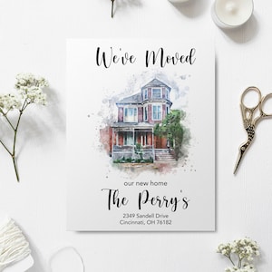 A "We’ve Moved" announcement card featuring a custom watercolor illustration of a two-story Victorian-style brick house. The card includes elegant script calligraphy reading "The Perry’s" and "We've Moved," with a placeholder address for Cincinnati, Ohio. The card is styled on a white wooden surface with baby's breath flowers, scissors, and twine.