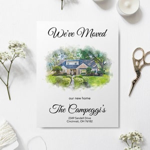 A vertical "We’ve Moved" announcement card featuring a custom watercolor portrait of a suburban house. The card includes elegant script text that reads "The Campeggi’s" and a sample address at the bottom. The card is styled on a white wooden surface surrounded by white baby's breath flowers, a spool of twine, and vintage gold scissors.
