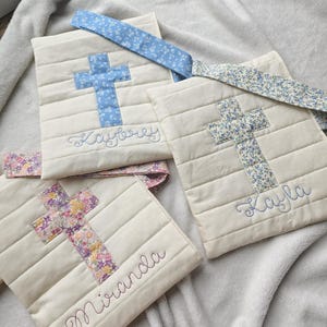 May include: Three white fabric bags with cross designs and personalized names. The bags have floral patterned straps. The names on the bags are Kaybree, Kayla, and Miranda.