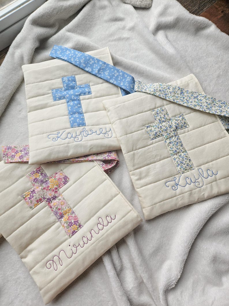 May include: Three white fabric bags with cross designs and personalized names. The bags have floral patterned straps. The names on the bags are Kaybree, Kayla, and Miranda.