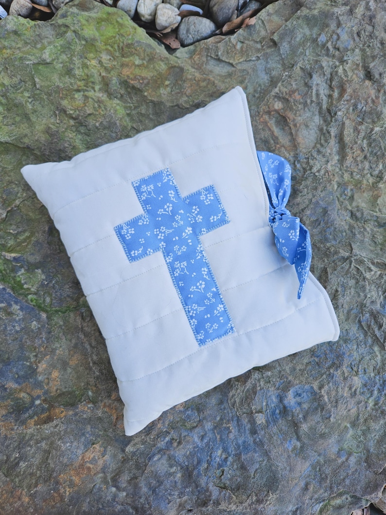 May include: A white quilted pillow with a blue cross design and a blue ribbon. The cross is made of fabric with a small white floral pattern.