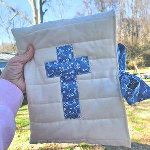 May include: A white quilted fabric pouch with a blue cross design. The cross is made of a blue fabric with a white floral pattern. The pouch has a blue ribbon tied around it.