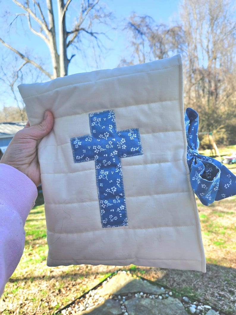 May include: A white quilted fabric pouch with a blue cross design. The cross is made of a blue fabric with a white floral pattern. The pouch has a blue ribbon tied around it.