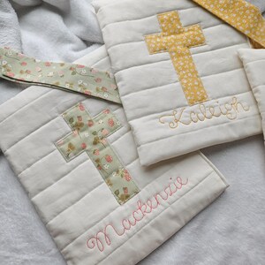May include: Two white quilted fabric bags with cross designs and embroidered names. The bag on the left has a green cross with a floral pattern and the name "Mackenzie" embroidered in pink. The bag on the right has a yellow cross with a floral pattern and the name "Kaleigh" embroidered in gold.