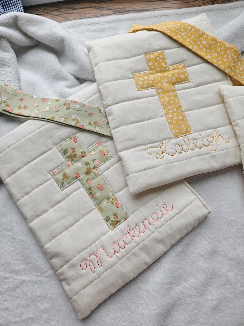 May include: Two white quilted fabric bags with cross designs and embroidered names. The bag on the left has a green cross with a floral pattern and the name "Mackenzie" embroidered in pink. The bag on the right has a yellow cross with a floral pattern and the name "Kaleigh" embroidered in gold.