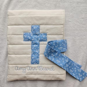 May include: A white quilted baby blanket with a blue cross and the name "Amy Ann Beard" embroidered on it. The blanket comes with a matching blue ribbon with a white floral pattern.
