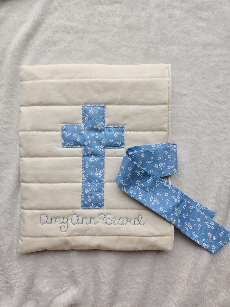 May include: A white quilted baby blanket with a blue cross and the name "Amy Ann Beard" embroidered on it. The blanket comes with a matching blue ribbon with a white floral pattern.