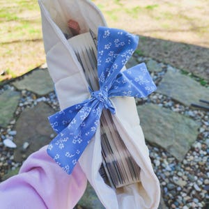 May include: A white fabric book sleeve with a blue and white floral ribbon tied around it. The sleeve is filled with a stack of books.