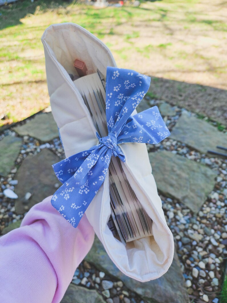 May include: A white fabric book sleeve with a blue and white floral ribbon tied around it. The sleeve is filled with a stack of books.