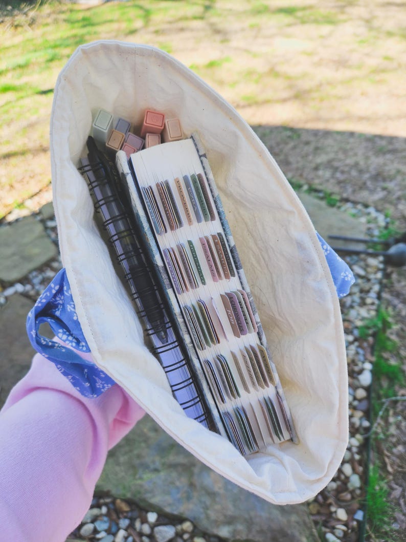 May include: A white canvas tote bag with blue floral trim, filled with a stack of notebooks, pens, and colorful markers.