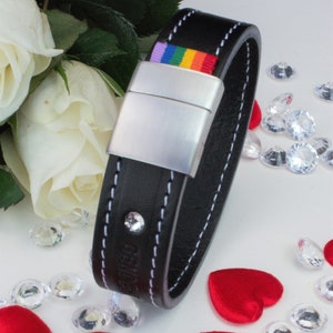 May include: Black leather bracelet with a rainbow stripe and silver clasp. The bracelet has white stitching and a small crystal. White roses and red heart-shaped confetti surround the bracelet. The bracelet is approximately 2 cm wide.