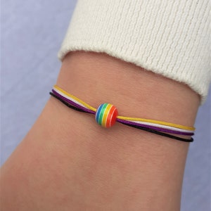 以下が含まれることがあります： 虹色のビーズと黒、白、紫、黄色の紐が付いたブレスレットです。