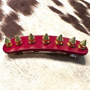 May include: A red leather hair clip adorned with seven gold-colored cone-shaped spikes. The hair accessory features a silver metal clip on the back. The clip is designed for securing hair and adds a bold, edgy style.