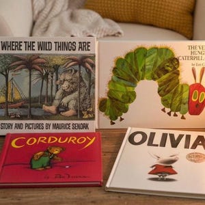 May include: Four children's books are displayed on a wooden surface. Titles include "Where the Wild Things Are," "The Very Hungry Caterpillar," "Corduroy," and "Olivia." A gold-colored lamp with a lit bulb is also present.