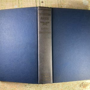 May include: A blue hardcover book with the title "The American Eagle" on the spine. The book is closed and the spine is visible. The book is on a wooden surface.