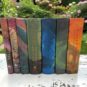 May include: A collection of seven Harry Potter books, each with a different colored spine. The books are arranged in a row on a white surface, with a blurred green and pink floral background. The titles and author's name are visible.