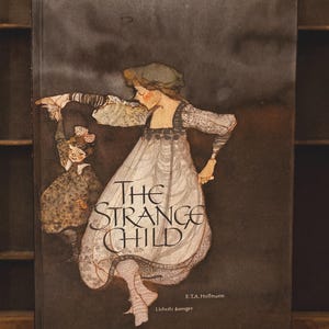 May include: A hardcover book titled "The Strange Child" with an illustration of two figures in vintage clothing. The cover is dark with the title in large, elegant lettering. The author's name is also visible.