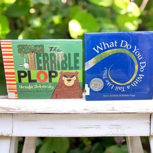 May include: Two children's books on a white wooden table. The book on the left is green with red and white stripes and features a bear and the title "The Terrible Plop". The book on the right is blue with a white spiral and the title "What Do You Do With a Tail Like This?"