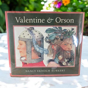May include: A book titled "Valentine & Orson" by Nancy Ekholm Burkert. The cover features an illustration of two people, one wearing a helmet with a feather and the other with a crown of leaves and a beard.