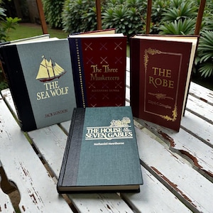 May include: A collection of hardcover books with titles including "The Sea Wolf," "The Three Musketeers," "The Robe," and "The House of the Seven Gables." The books have various colors, including teal, burgundy, and dark gray.