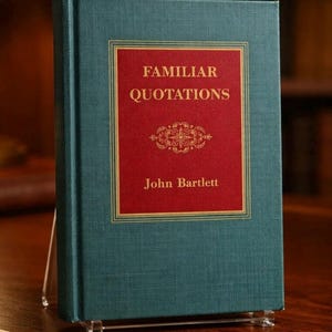 May include: A hardcover book titled "Familiar Quotations" by John Bartlett. The book has a teal cover with a red rectangular inset featuring gold lettering and decorative elements. The book is propped up on a clear acrylic stand.
