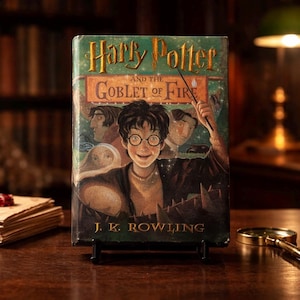 May include: A hardcover copy of "Harry Potter and the Goblet of Fire" by J.K. Rowling. The book cover features a smiling illustration of Harry Potter, with the title in gold lettering. The book is displayed on a black stand.