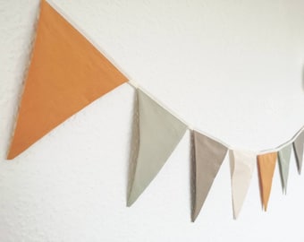 Könnte beinhalten: Eine Girlande aus Stofffahnen in verschiedenen Brauntönen, Beige und Orange. Die Fahnen sind mit weißem Garn zusammengefügt und hängen an einer weißen Wand.