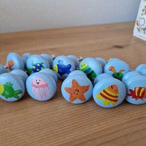 Könnte beinhalten: Eine Sammlung hellblauer Holzschrankknöpfe, die jeweils mit einem anderen Meereskreaturen-Design handbemalt sind. Die Knöpfe zeigen eine Vielzahl farbenfroher Meereslebewesen, darunter eine Krabbe, ein Fisch, ein Seestern und eine Qualle. Die Knöpfe liegen auf einer Holzoberfläche.