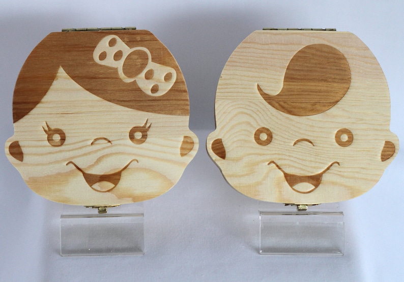 Puede incluir: Dos cajas de dientes de madera con un dise&ntilde;o de cara de beb&eacute; sonriente. Una caja tiene una ni&ntilde;a con un lazo en la cabeza y la otra caja tiene un ni&ntilde;o con pelo. Ambas cajas tienen un soporte de acr&iacute;lico transparente.
