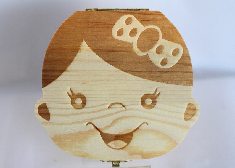 Puede incluir: Caja de madera para el hada de los dientes con un rostro de ni&ntilde;a tallado y un lazo en la cabeza. La caja es de color marr&oacute;n claro con veta de madera natural.