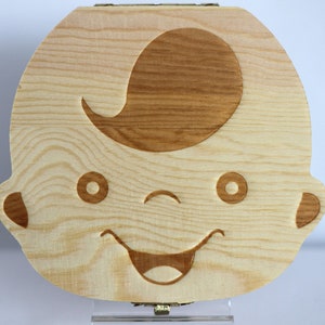 Puede incluir: Una caja de dientes de hada de madera con forma de cara de ni&ntilde;o con una boca sonriente y ojos cerrados. La caja est&aacute; hecha de madera marr&oacute;n claro con un dise&ntilde;o de madera marr&oacute;n oscuro para el cabello y los rasgos faciales.
