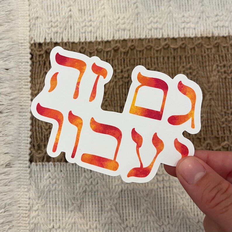 May include: White sticker with Hebrew text in orange and red gradient. The sticker is held by a hand against a textured background. The text appears to be a phrase or saying.